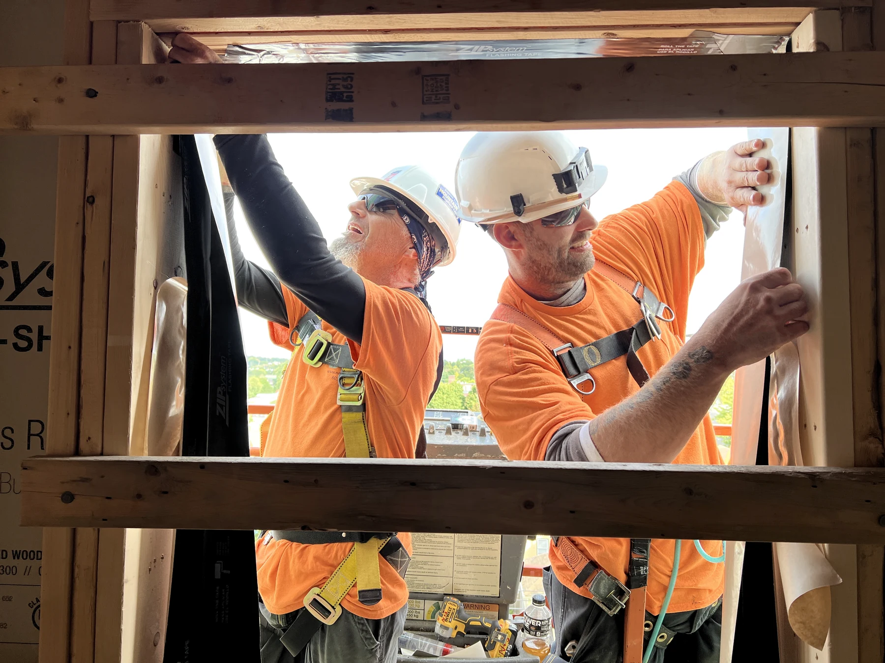 Two carpenters in orange shirts and white hard hats working together around a framed window opening, applying flashing tape and sheathing